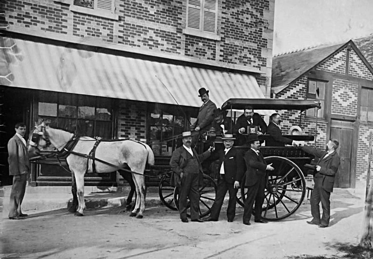 Photographie ancienne : un attelage et un groupe d’hommes devant la façade à store rayé de l’hôtel Baudy, à Giverny