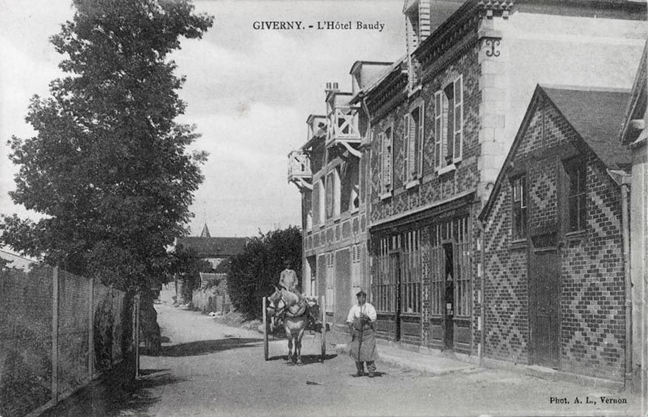 Carte postale ancienne : la façade de brique de l’hôtel Baudy vue depuis la rue, avec une charrette tirée par un âne